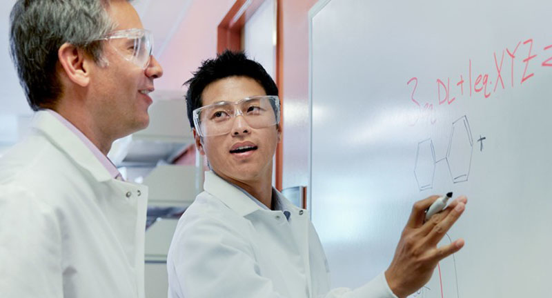 two men in lab coats and goggles. One facing the other and writing on a whiteboard.