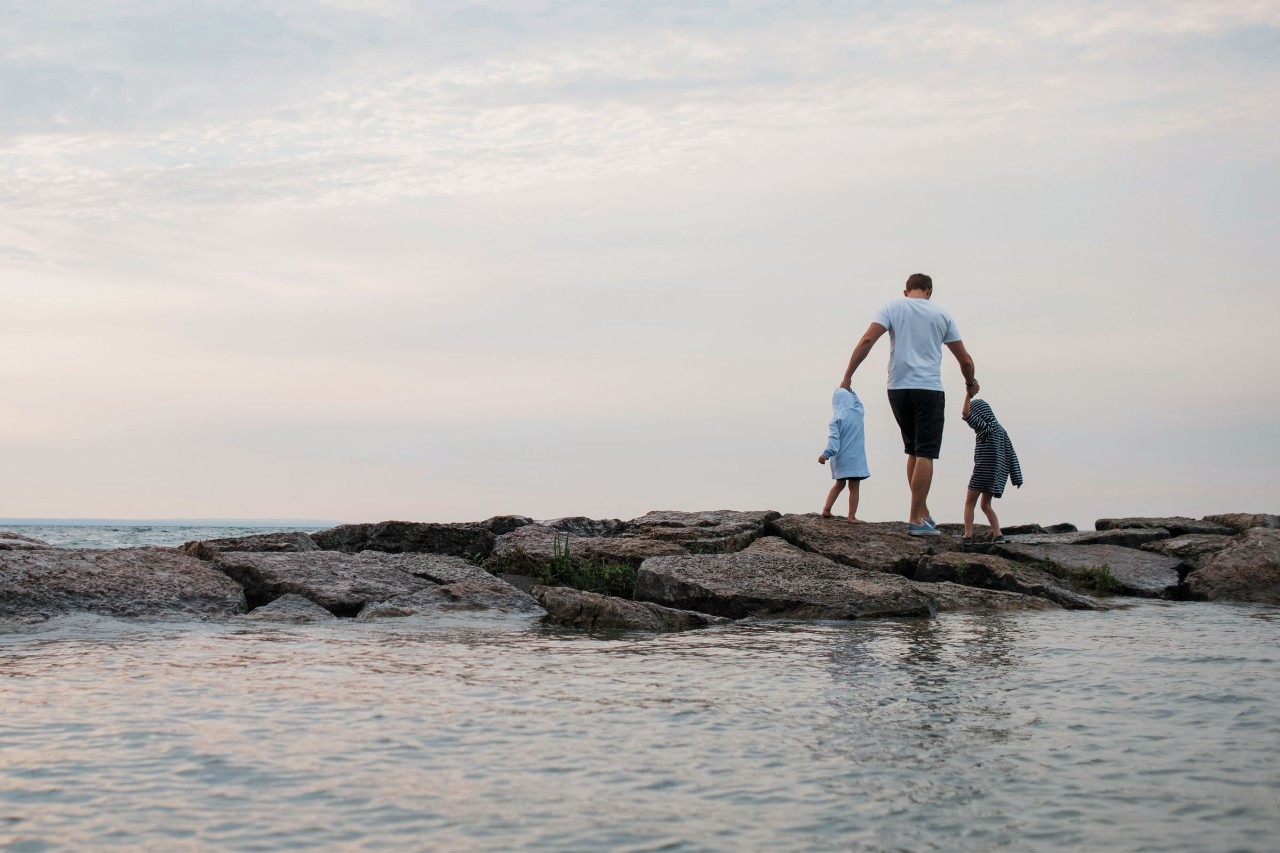 Man holding his kids hands while walking on rocks at the beach