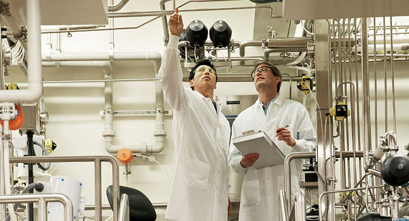 Two male scientists in lab coats pointing up toward the ceiling holding a notebook.